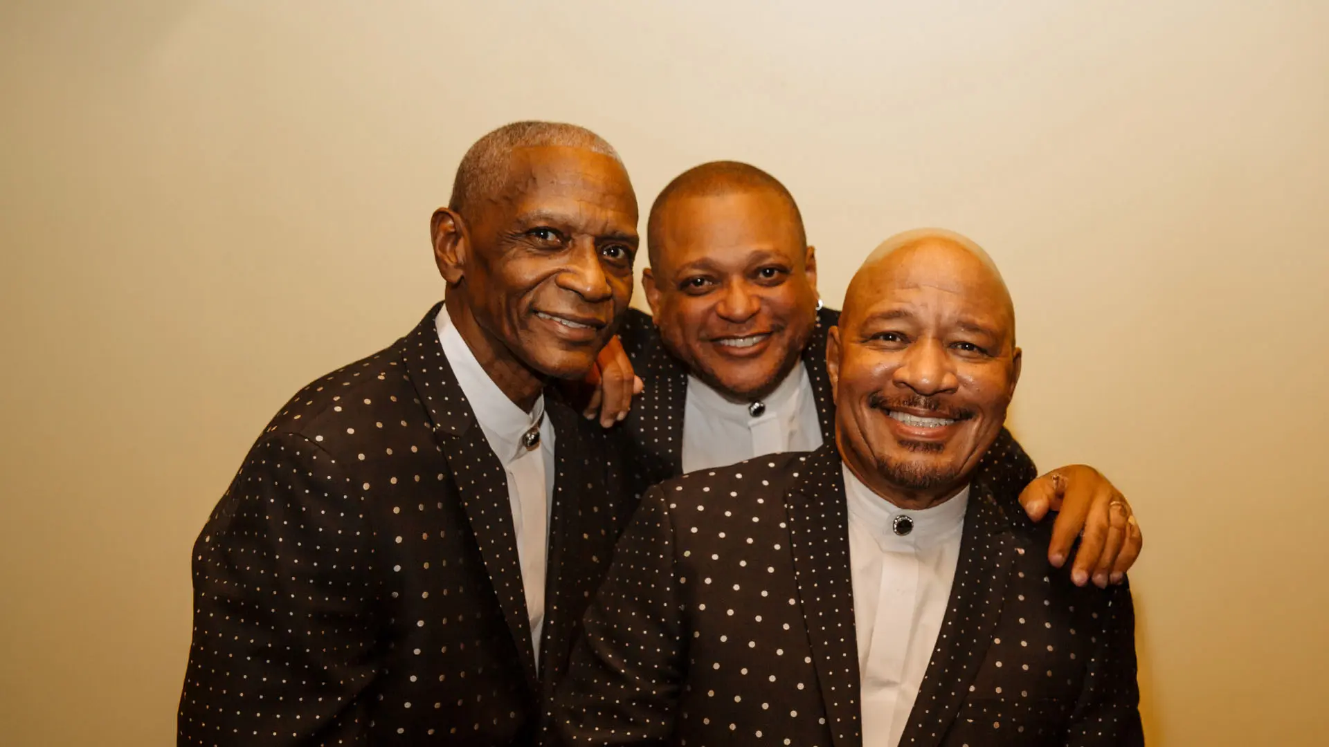 Three men stand closely together, all dressed in matching, polka-dotted black suits with white shirts. Each of them has a warm, engaging smile, suggesting a cheerful and friendly atmosphere. Their coordinating attire and joyful demeanor imply a sense of unity and camaraderie, possibly representing a musical group.