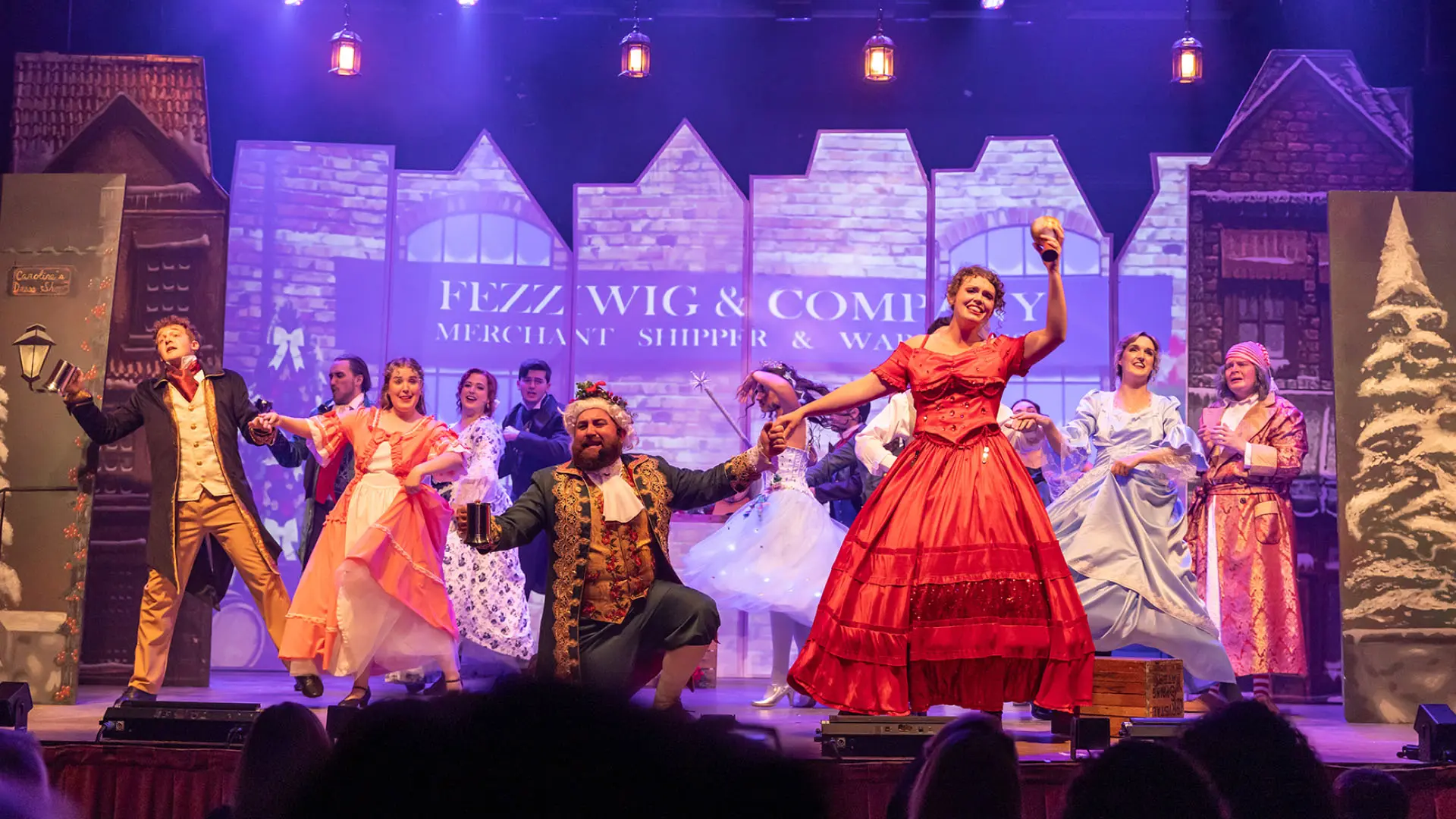 A lively stage performance featuring actors in elaborate Victorian costumes dancing energetically against a backdrop depicting an old-fashioned city street and a sign reading 'FEZZ WIG & COMPANY.' The scene is illuminated by bright stage lights, creating a warm and festive atmosphere, evoking the holiday spirit and the joy of traditional celebration.