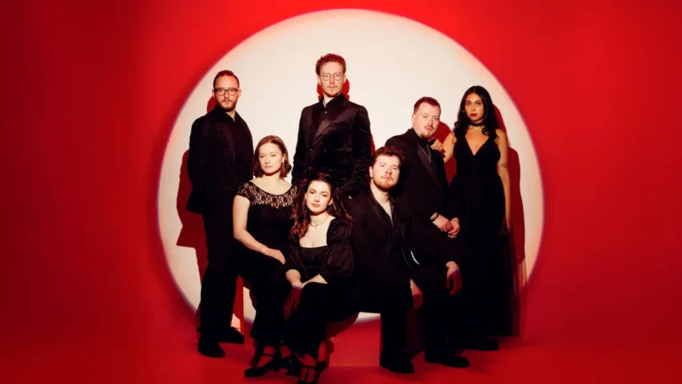 A well-dressed group poses elegantly against a vibrant red backdrop, creating a striking contrast. The individuals are wearing formal black attire, suggesting a sense of sophistication and unity. The composition is artistically lit, emphasizing the group's confident and cohesive appearance.