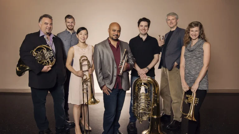 A group of seven musicians posing with brass instruments such as horns, trombones, and tubas. They stand in a relaxed manner, displaying a mix of formal and casual attire, reflecting a professional yet approachable music group.