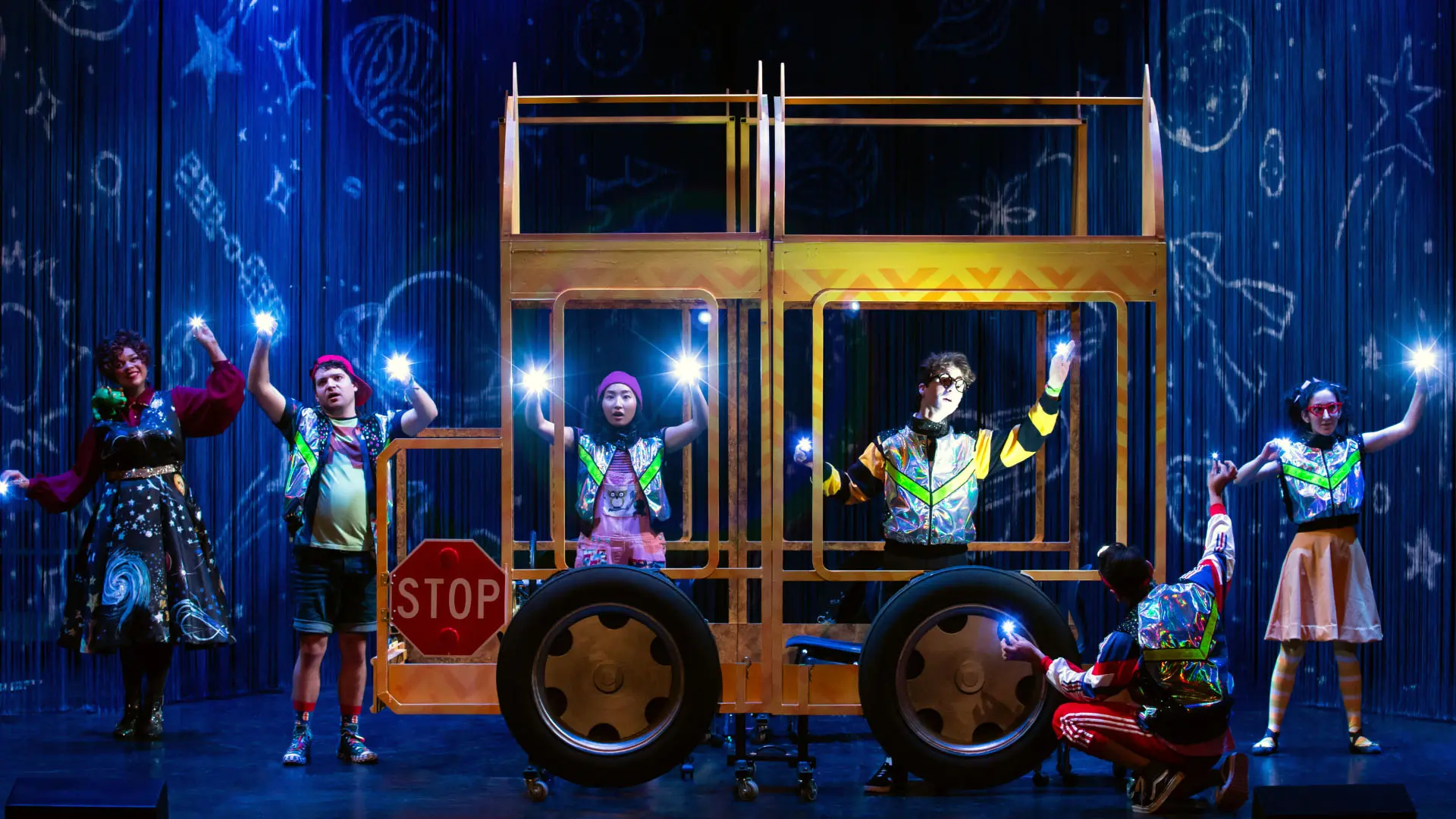 Performers are displayed in vibrant, eccentric costumes, standing on a stage featuring a bus set piece. The backdrop is illustrated with cosmic and whimsical drawings such as stars and planets, creating an imaginative atmosphere. Each actor holds a glowing light, contributing to the playful and fantastical theme of the performance.