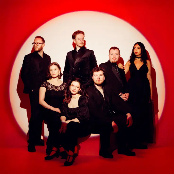 A well-dressed group poses elegantly against a vibrant red backdrop, creating a striking contrast. The individuals are wearing formal black attire, suggesting a sense of sophistication and unity. The composition is artistically lit, emphasizing the group's confident and cohesive appearance.