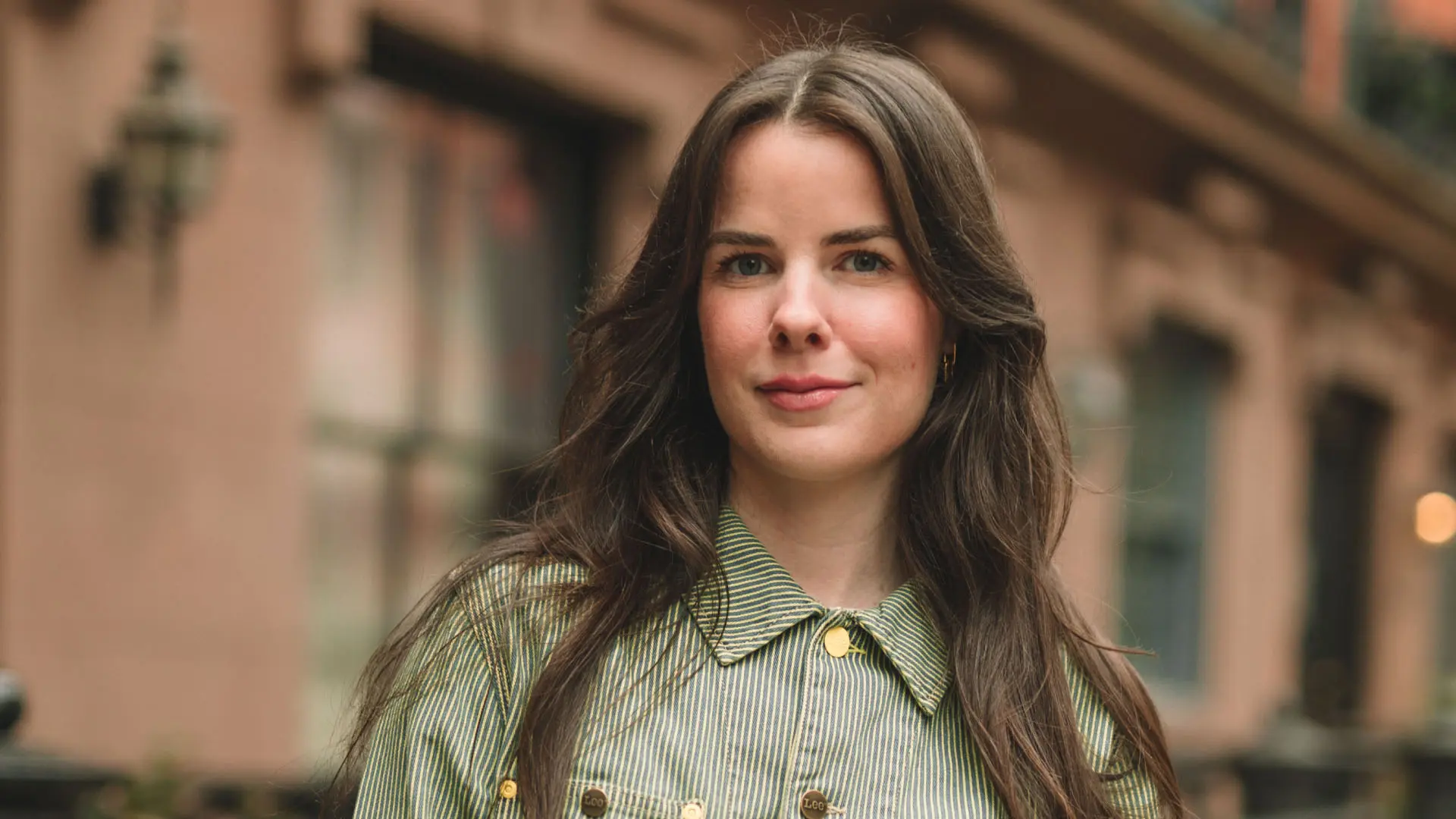 A young woman with long brown hair and a relaxed expression stands against an urban backdrop, wearing a striped green jacket. The architecture in the background hints at a city street setting