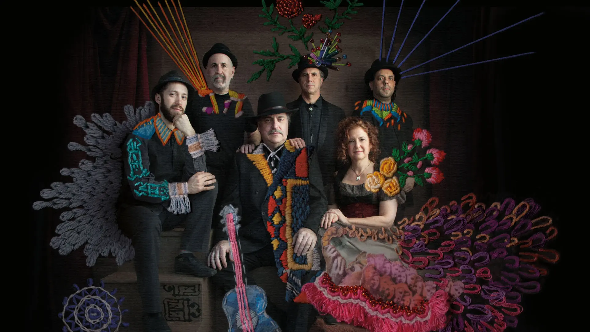 A group of six individuals, dressed in dark attire, are seated and standing in a formal arrangement. Each person's outfit is uniquely embellished with vibrant, embroidered patterns and textures, including floral motifs and geometric designs. These embroidered elements extend into the surrounding space, creating an artistic and imaginative visual effect.