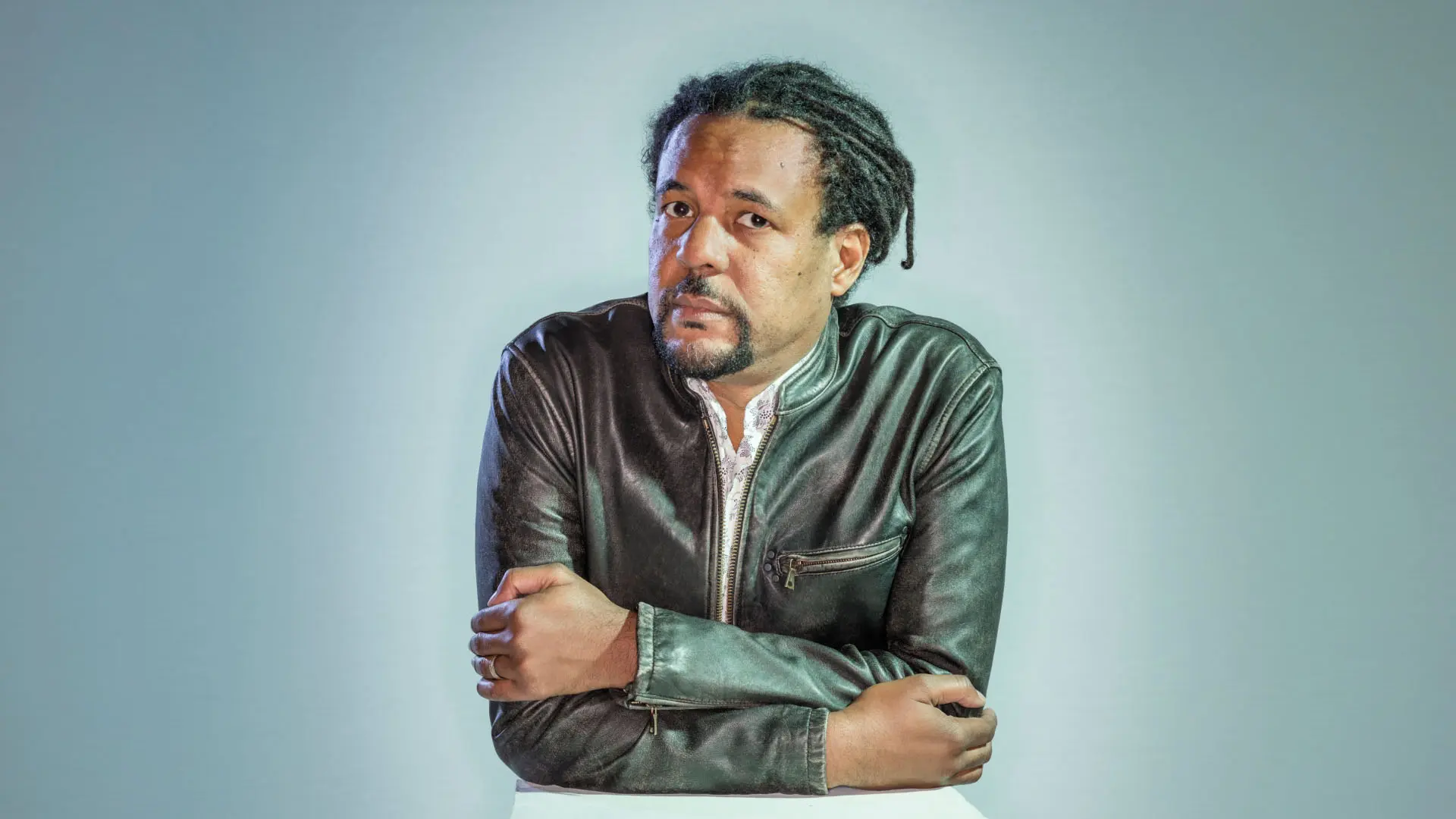 A man with a thoughtful expression poses with his arms crossed, wearing a stylish leather jacket. His hair is styled in dreadlocks, and the background is a soft gradient.