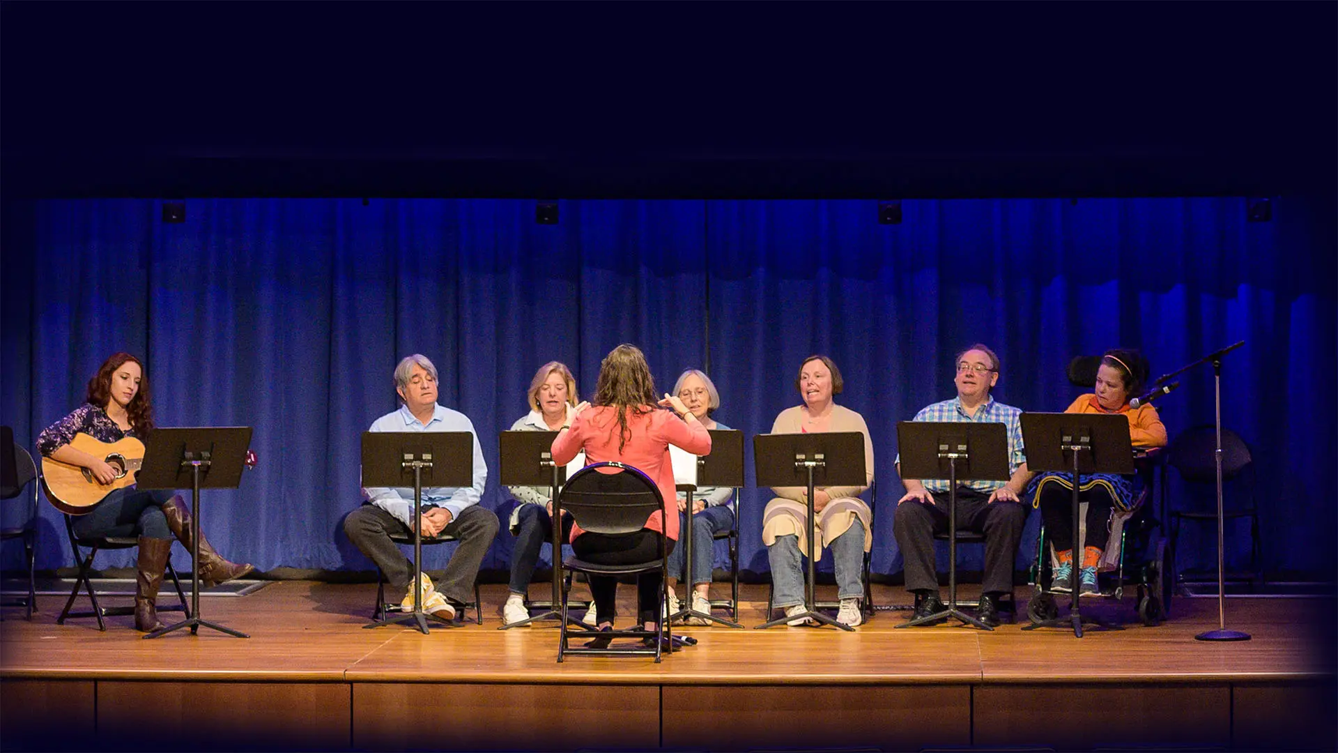 A group of adults sits in chairs on a stage in front of music stands. One person to the right is in a wheelchair. They are focused on a person standing in front of them. One person plays the guitar, accompanying the group. The scene is set against a blue backdrop and lit with warm stage lights, creating an inviting atmosphere.