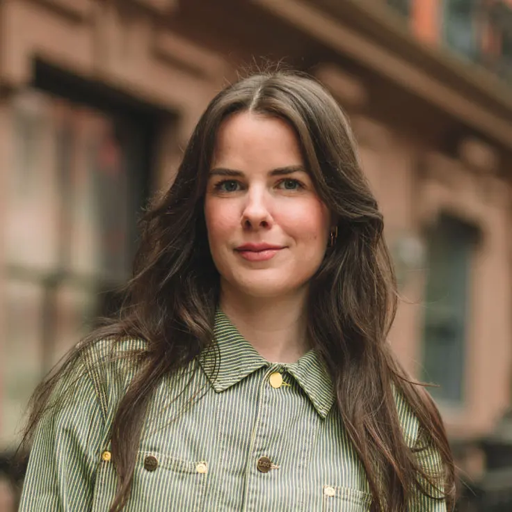 A young woman with long brown hair and a relaxed expression stands against an urban backdrop, wearing a striped green jacket. The architecture in the background hints at a city street setting