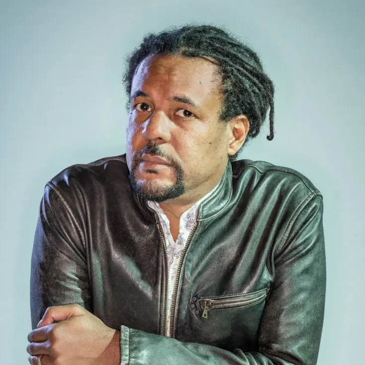 A man with a thoughtful expression poses with his arms crossed, wearing a stylish leather jacket. His hair is styled in dreadlocks, and the background is a soft gradient.