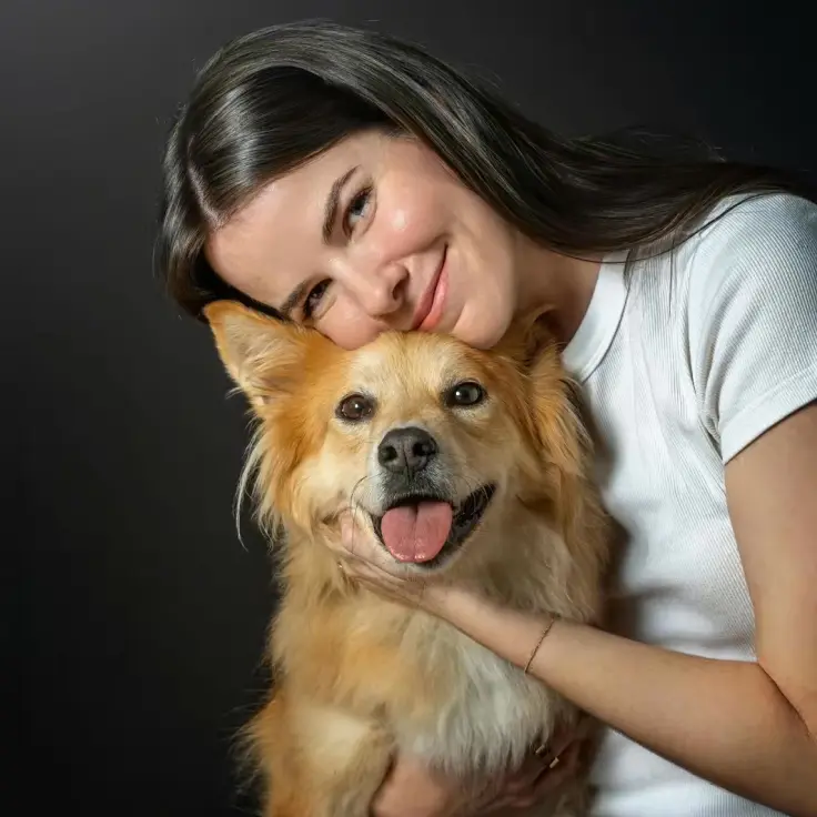 Isabel Klee in a white shirt cuddles a fluffy tan dog in a studio portrait against a dark background.