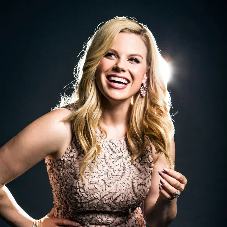 A woman with long blonde hair smiles brightly, exuding confidence and joy. She wears a sleeveless, patterned dress with subtle floral motifs, complemented by sparkling earrings.
